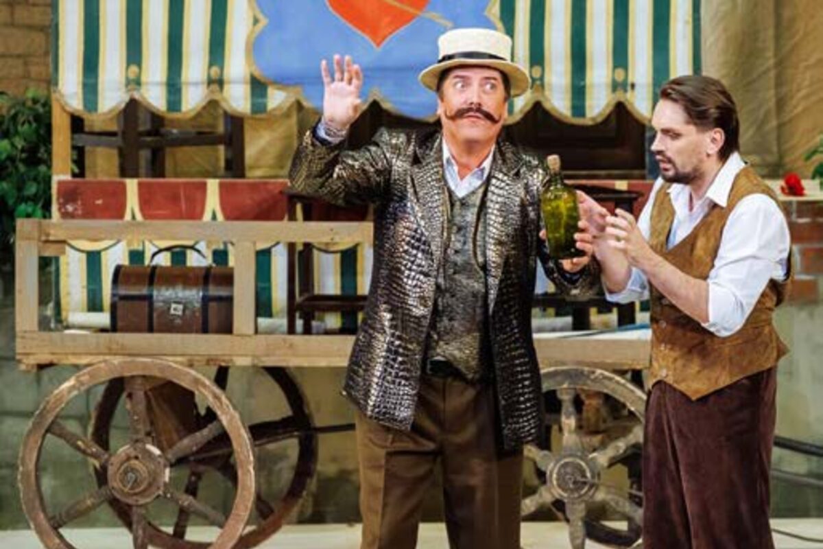 El charlatán Dulcamara, con chaqueta metalizada y sombrero de paja, ofrece un frasco de elixir a Nemorino frente a su carromato en L'Elisir d'Amore.
