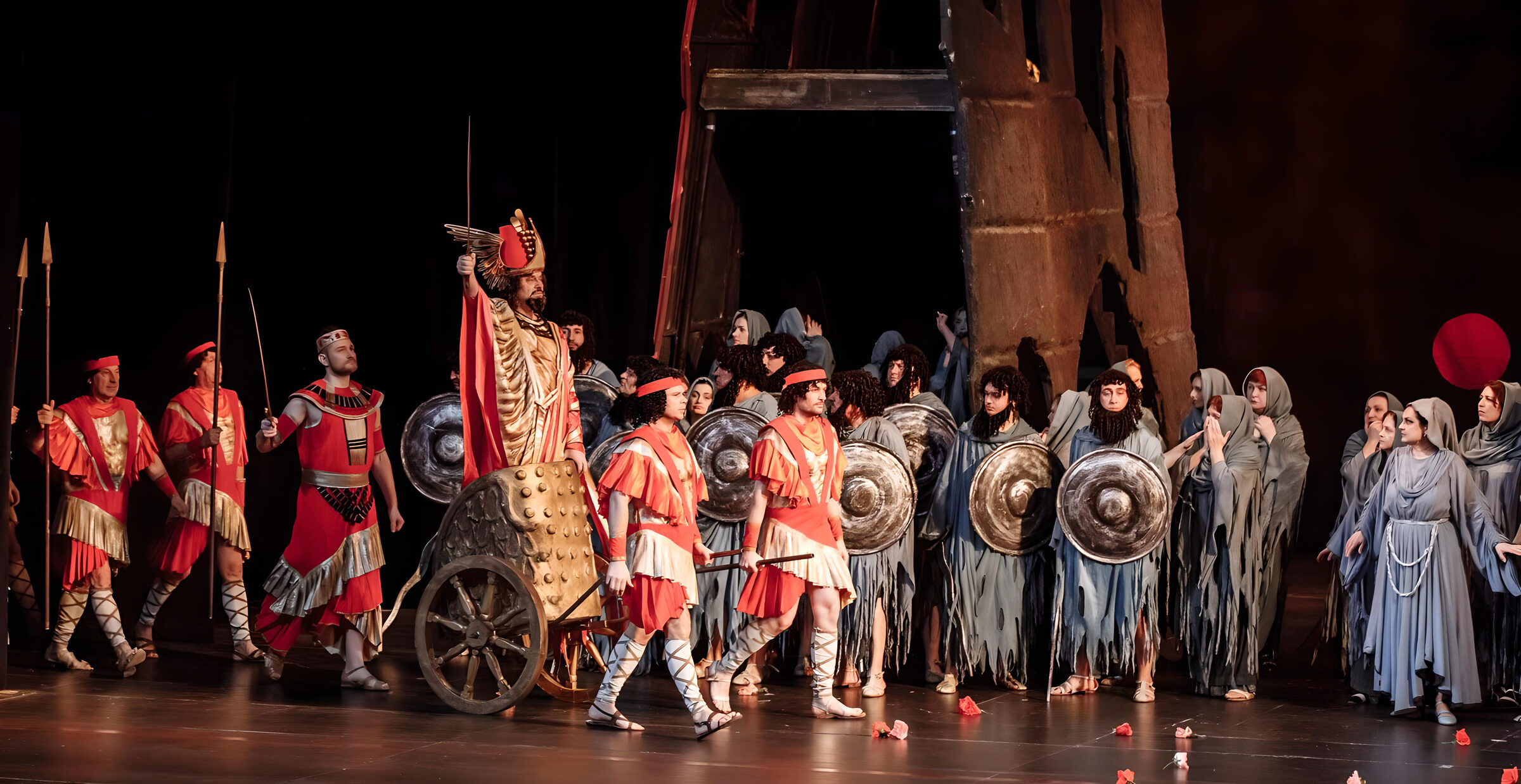 Escena coral de Nabucco: soldados con túnicas rojas y escudos junto a un carro dorado, y un coro de figuras en gris al fondo, sobre el escenario de la Ópera Nacional de Moldavia.