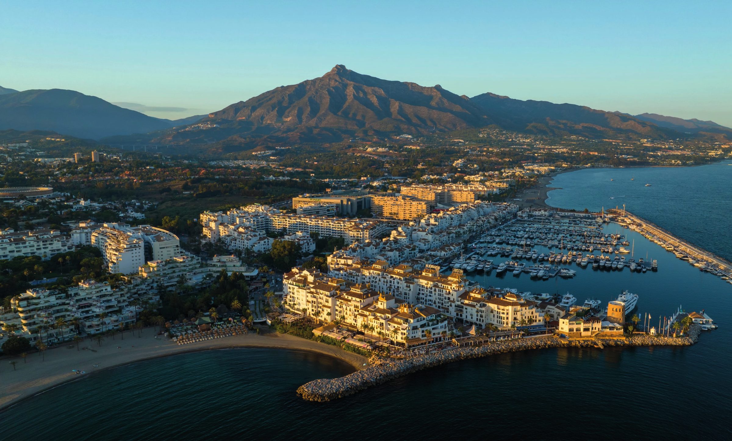 Vista aérea de Puerto Banús al atardecer, con el puerto deportivo, la playa y La Concha al fondo.